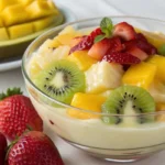 A bowl of Nata Fruit Pudding topped with fresh kiwi, mango, pineapple, and strawberries, with a background of sliced mango.
